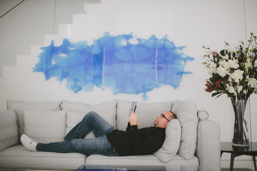 Shot of a man laying down and checking his tablet on a gray couch with a white wall in the background with blue artwork on it.