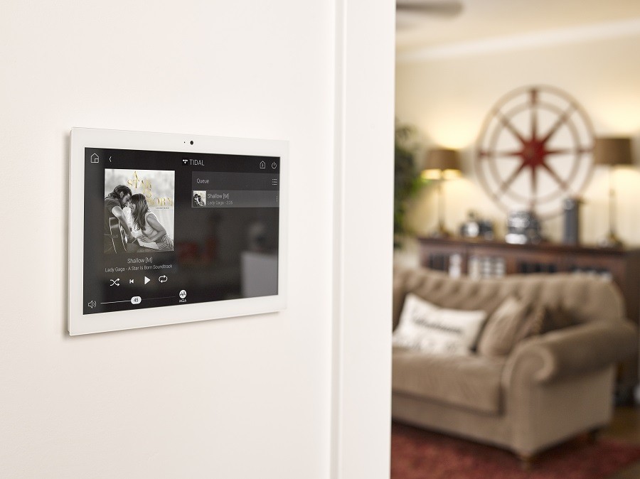 A Control4 touch panel on the wall shows an album cover with a living room in the background. 