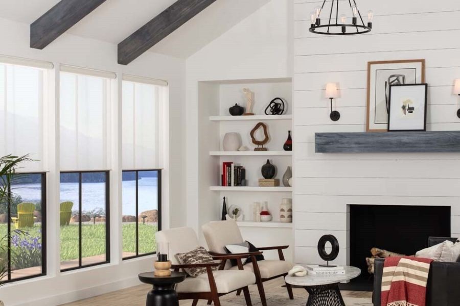 Photo of a cozy living room, white shiplap behind the fireplace mantel, large windows with motorized shades to the left, and the view of a lake beyond.
