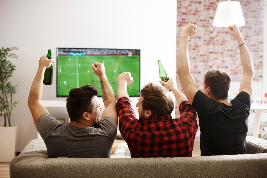 Three men sitting on a couch cheering on a soccer game on the TV.