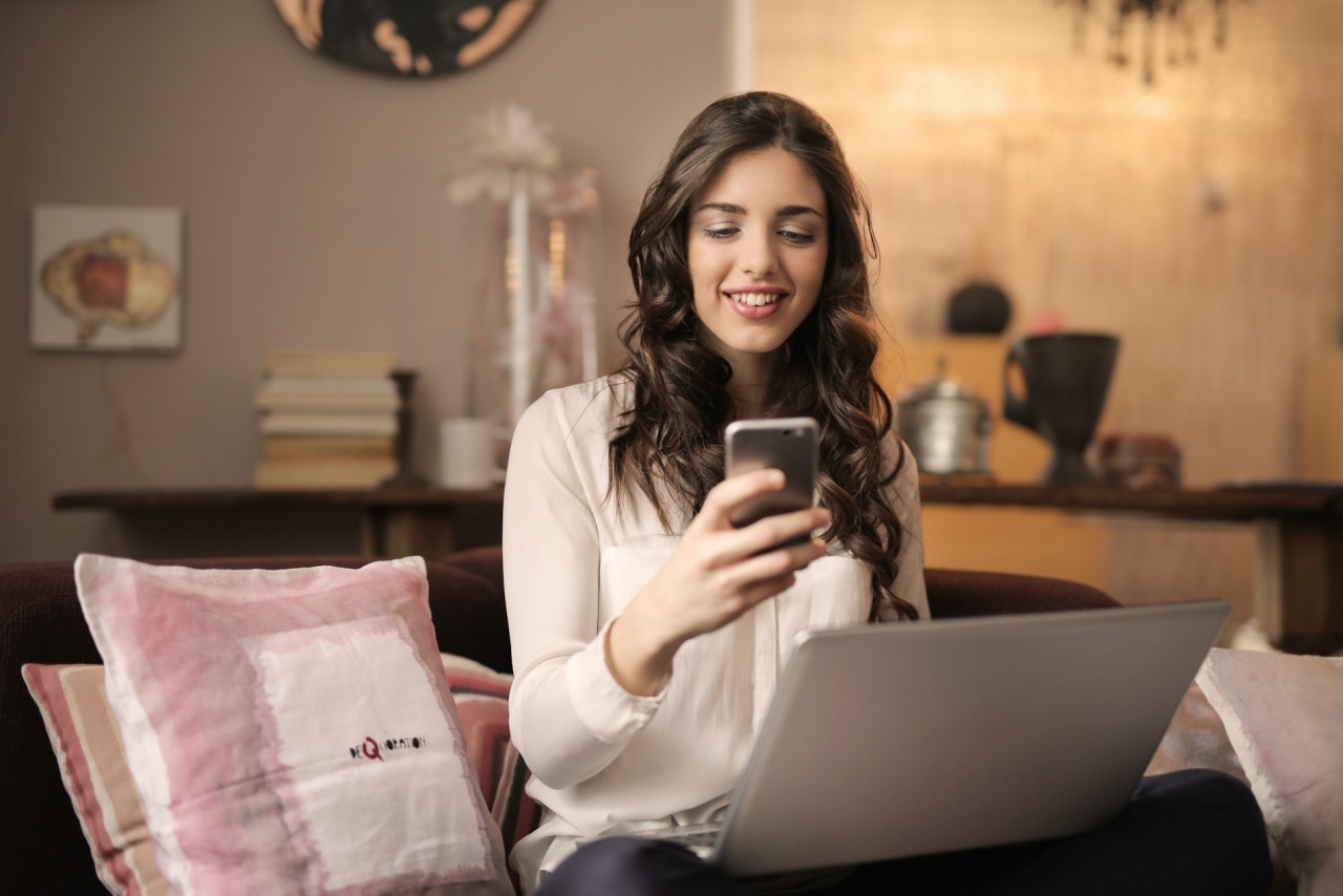 Woman sitting on her couch looking at her phone, a laptop open on her lap.