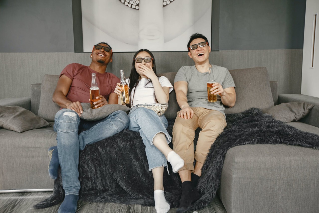 Two men and a woman sitting on a comfortable couch, laughing and eating popcorn.