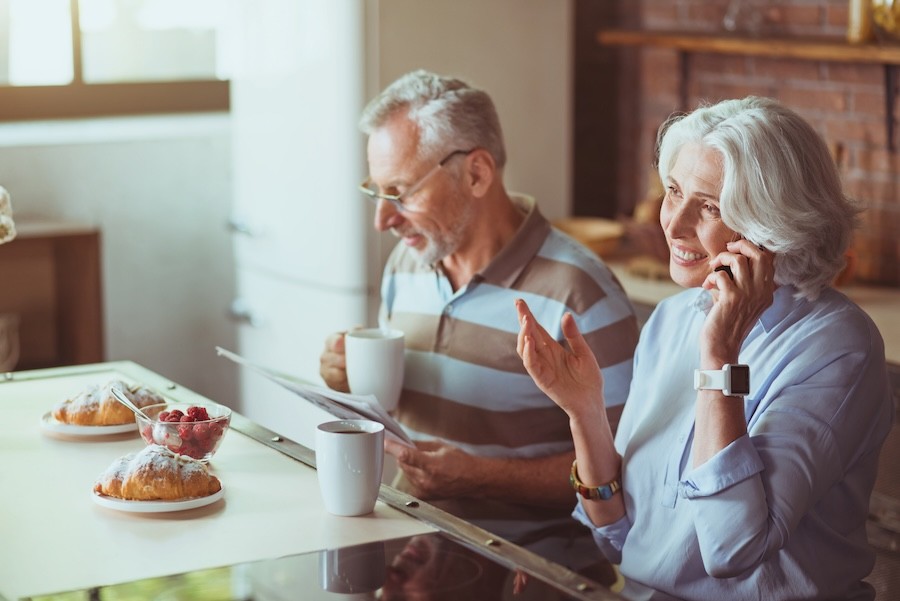 An aging couple sits at the kitchen table using devices.