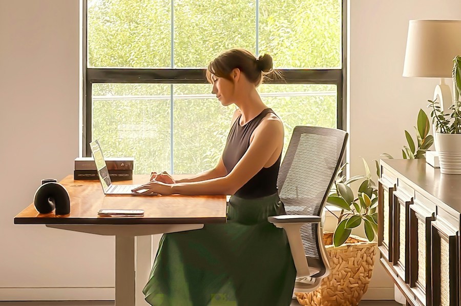 A woman works on a laptop at a desk near a window in her home office.